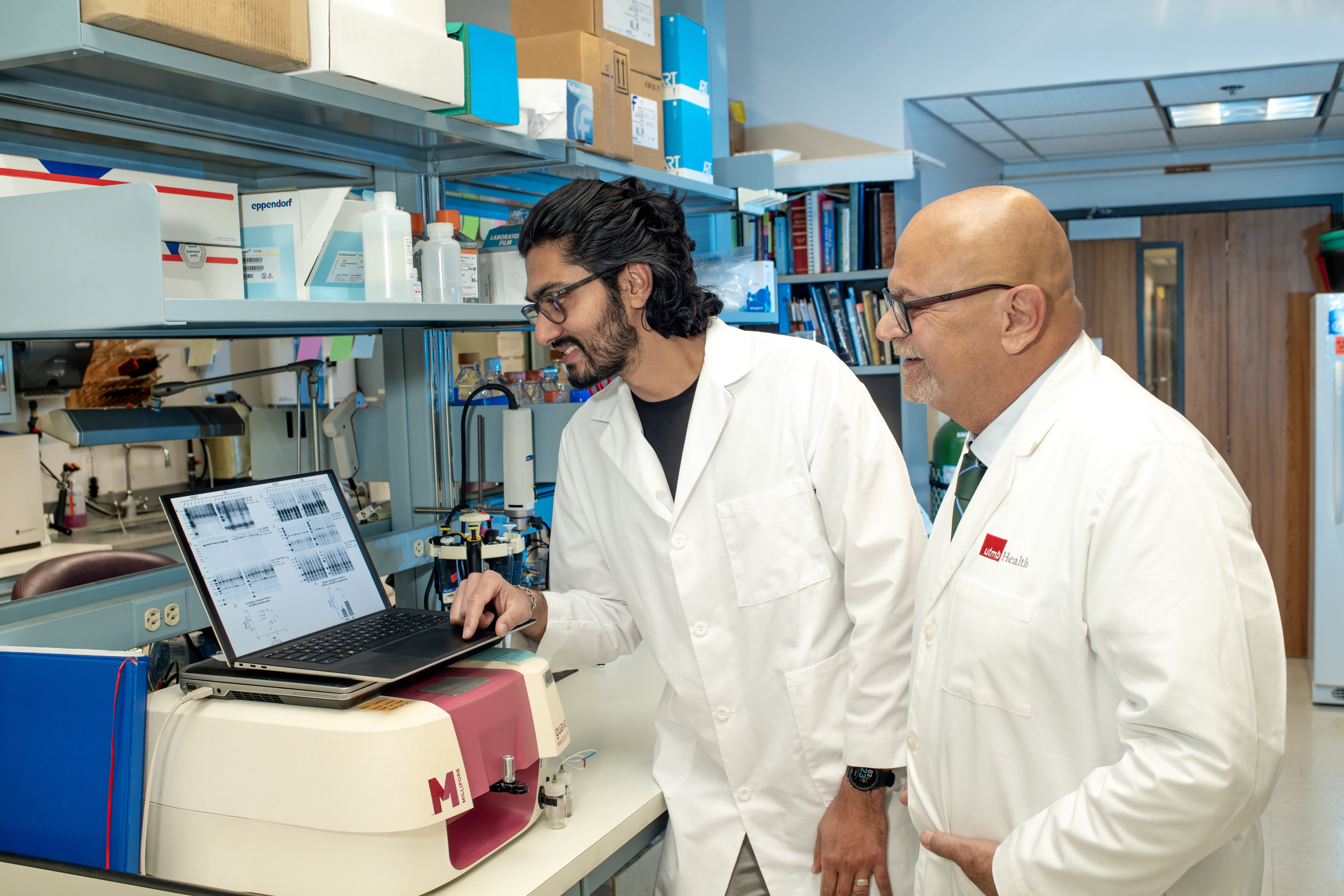 Two males wearing white coats standing in front of and looking at a laptop in a lab. they are both smiling