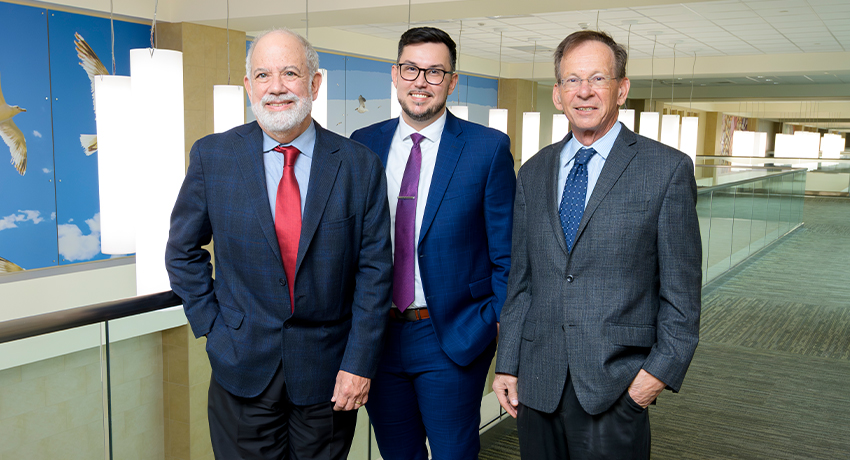 Three people in business suits stand side by side in a brightly lit indoor hallway with large hanging light fixtures. A wall featuring bird illustrations is visible behind them.