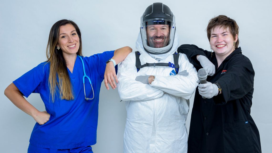 Three people stand side by side in different types of lab and medical attire, including scrubs, a protective suit, and a lab coat, with one person holding a microphone.