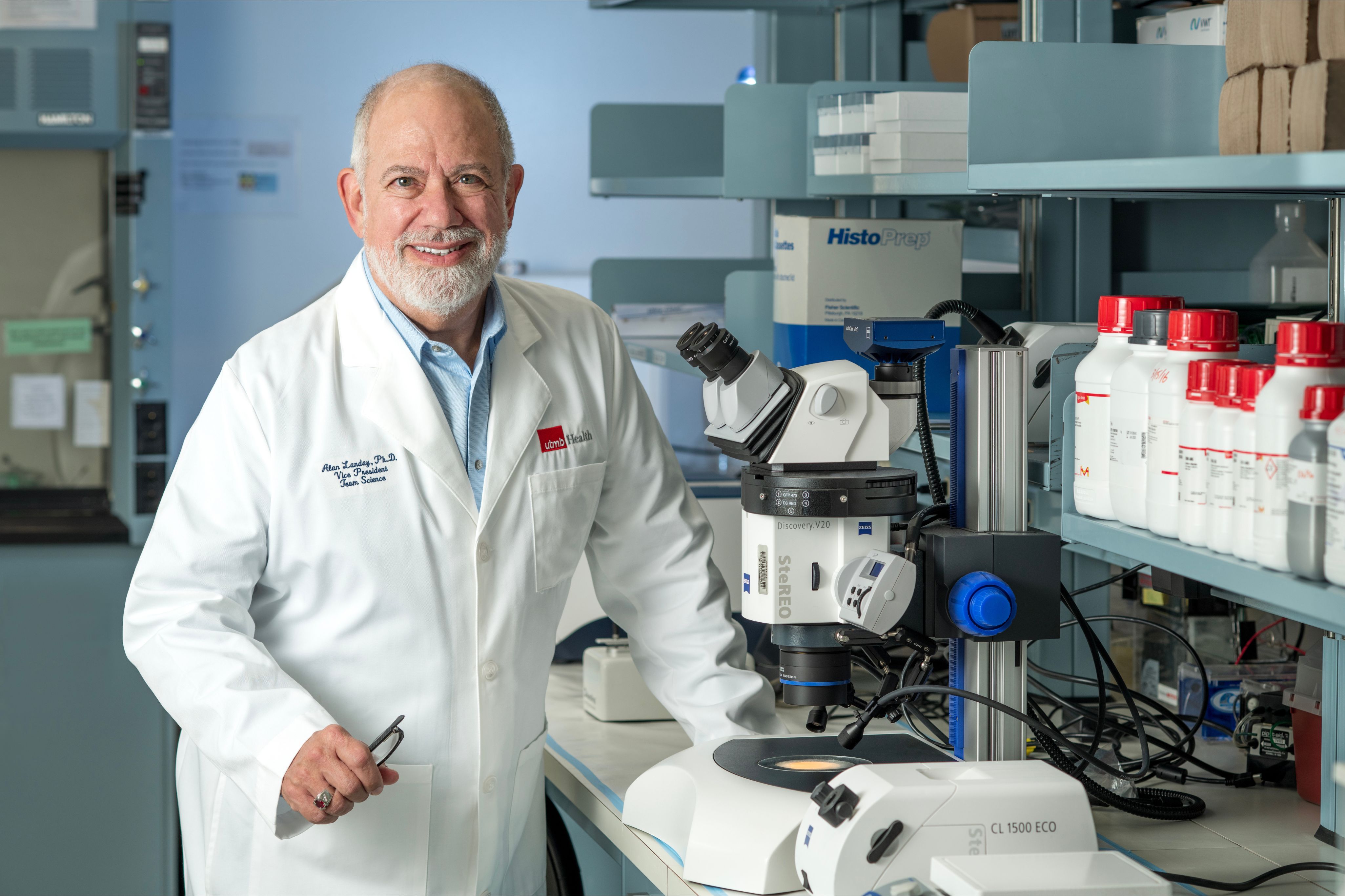 A person in a lab coat stands at a laboratory workstation beside a microscope and rows of labeled chemical bottles.