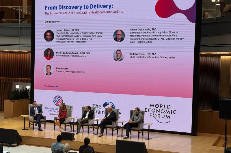 Panelists sit on a stage at the Brain Capital summit in Houston, speaking in front of a large screen titled “From Discovery to Delivery: The Economic Value of Accelerating Healthcare Innovations,” with an audience in the foreground