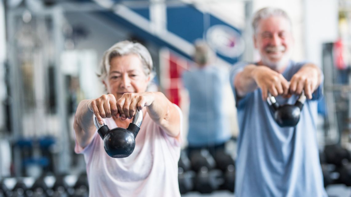 Two people in a gym hold kettlebells extended forward during a strength‑training exercise.