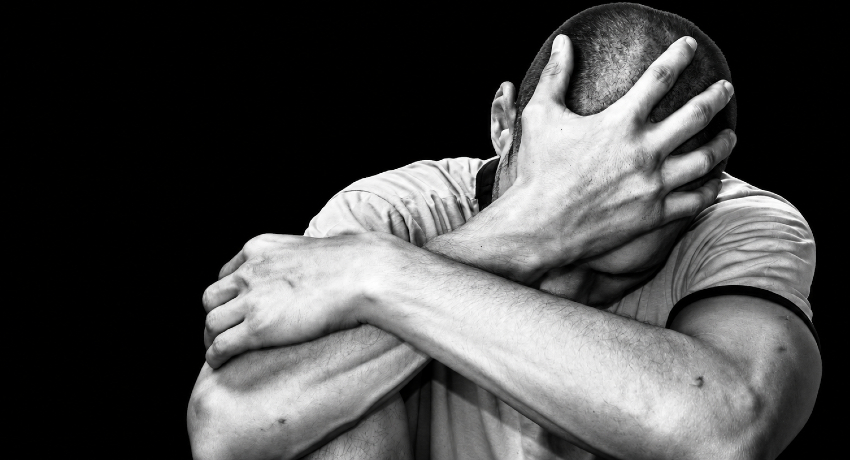 A man is shown seated with knees drawn up, one arm wrapped around his legs and a hand covering part of his head against a dark background.