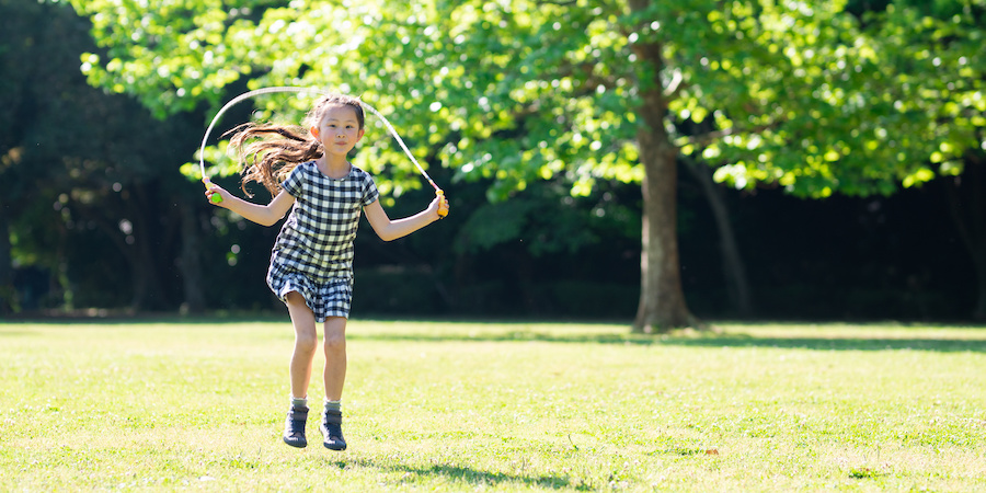 young brunette girl wearing a black and white checkered outfit jumps rope on grass with a tree in the background